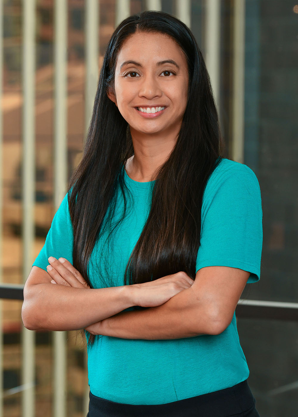 A woman with long dark hair, wearing a teal short-sleeve shirt, stands indoors in front of large windows with vertical blinds, smiling with her arms crossed.