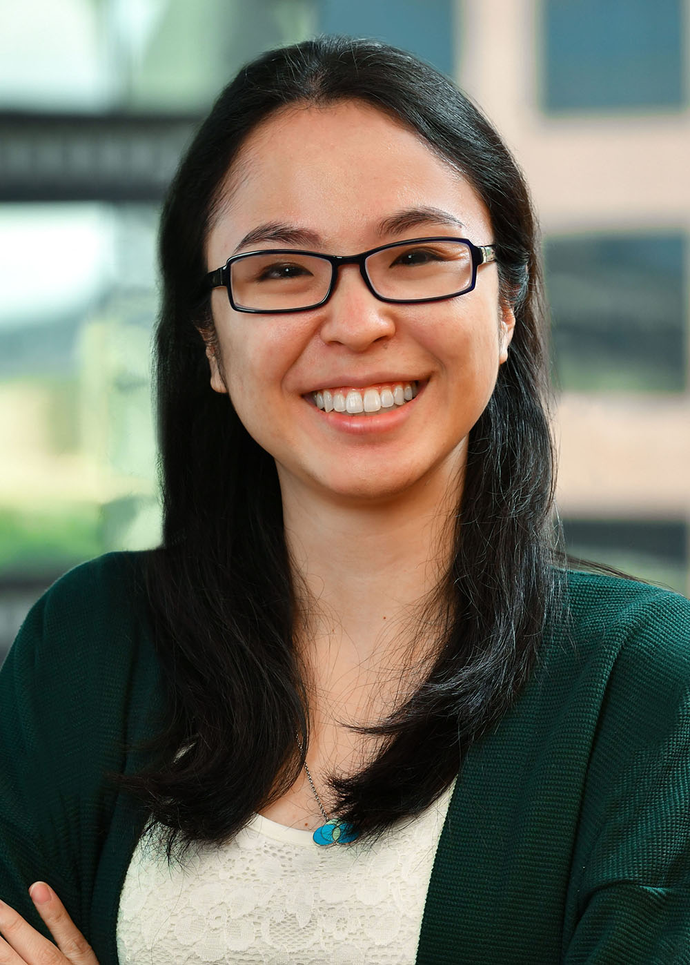 A woman with black hair and glasses smiles, wearing a green cardigan over a white lace top.