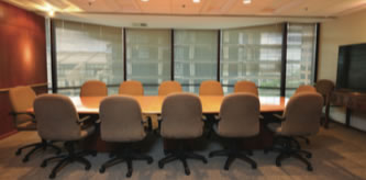 A conference room with a long wooden table surrounded by ten beige office chairs, large windows with blinds, and a flat-screen TV on the right wall.