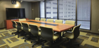 A modern conference room with a long wooden table surrounded by ten padded chairs. Large windows offer a view of tall office buildings outside. A flat-screen TV is mounted on a cabinet in the corner.