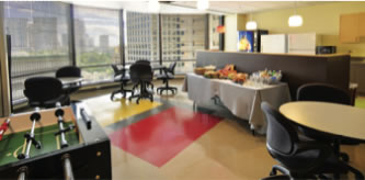 Modern office break room with large windows, round tables and chairs, a foosball table, and a buffet table with snacks and drinks. City buildings are visible outside the windows.