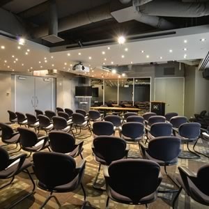 Rows of empty black chairs are arranged facing a presentation area in a modern conference room with overhead lighting and exposed ductwork.