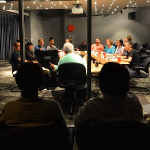 A group of people sit around a conference table having a meeting in a dimly lit room, while two people watch from behind a glass window.