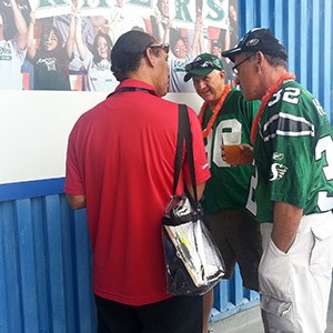 Three men stand and chat near a wall. Two wear green football jerseys and leis, holding drinks, while the third, seen from the back, wears a red shirt and carries a clear bag. A crowd is visible in a poster behind them.