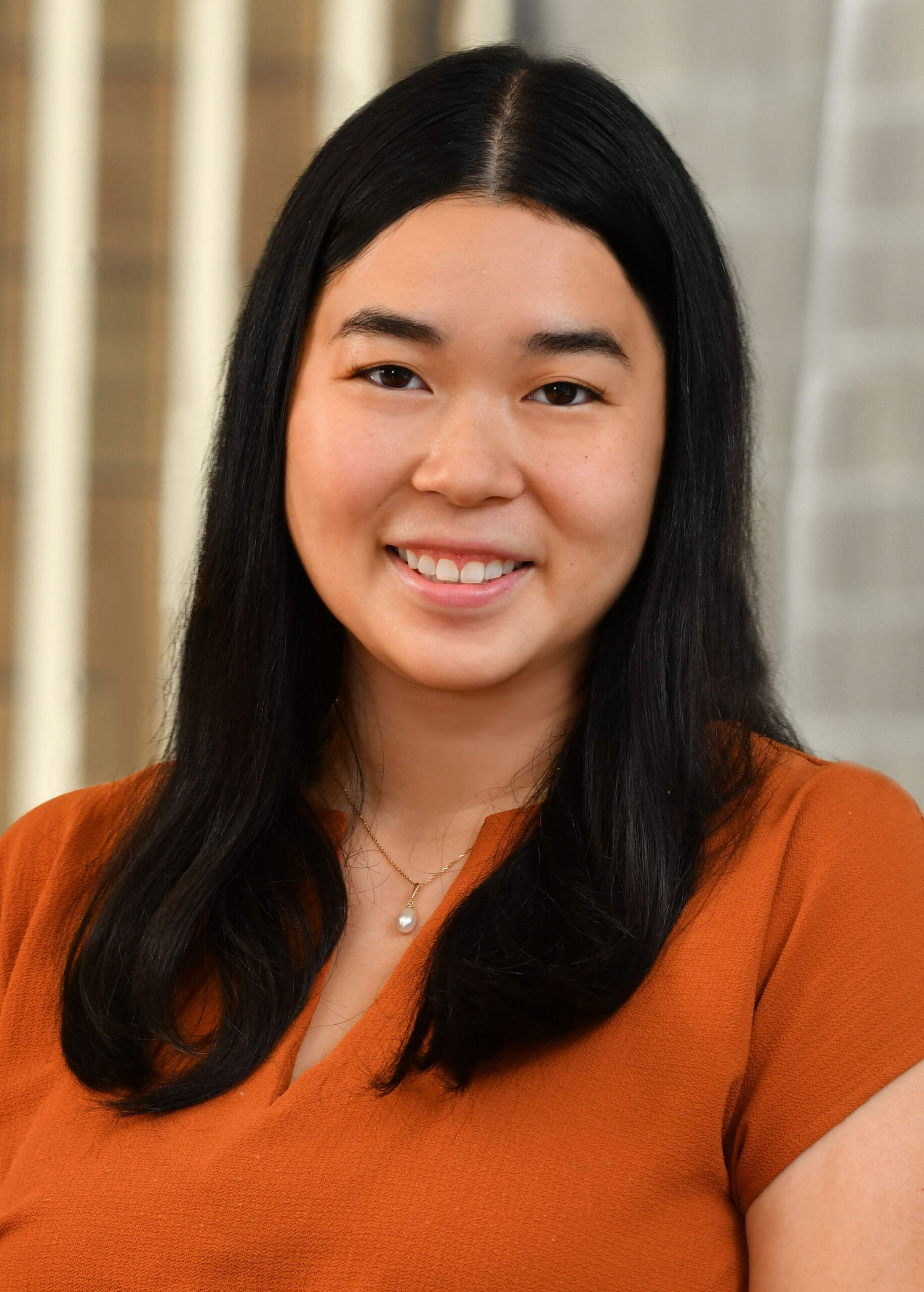 A woman with long black hair, wearing an orange top and a necklace, smiles at the camera against a blurred indoor background.