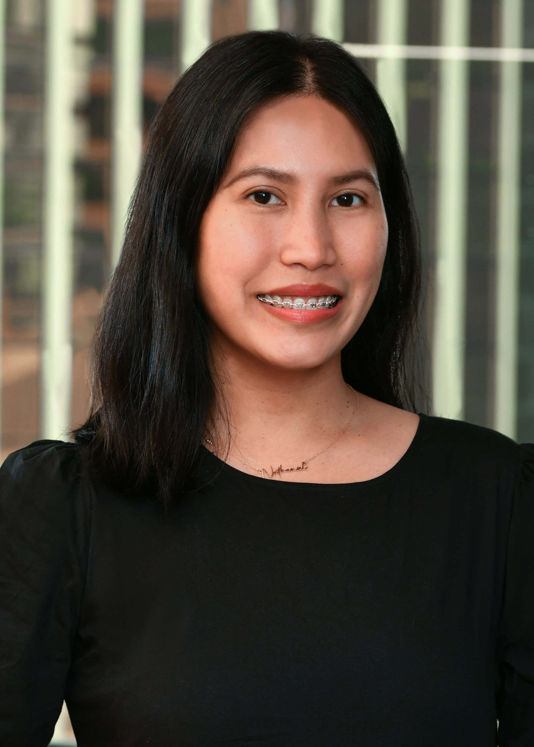 A woman with long dark hair, wearing a black top and braces, smiles at the camera. Vertical blinds and a softly blurred background are visible behind her.