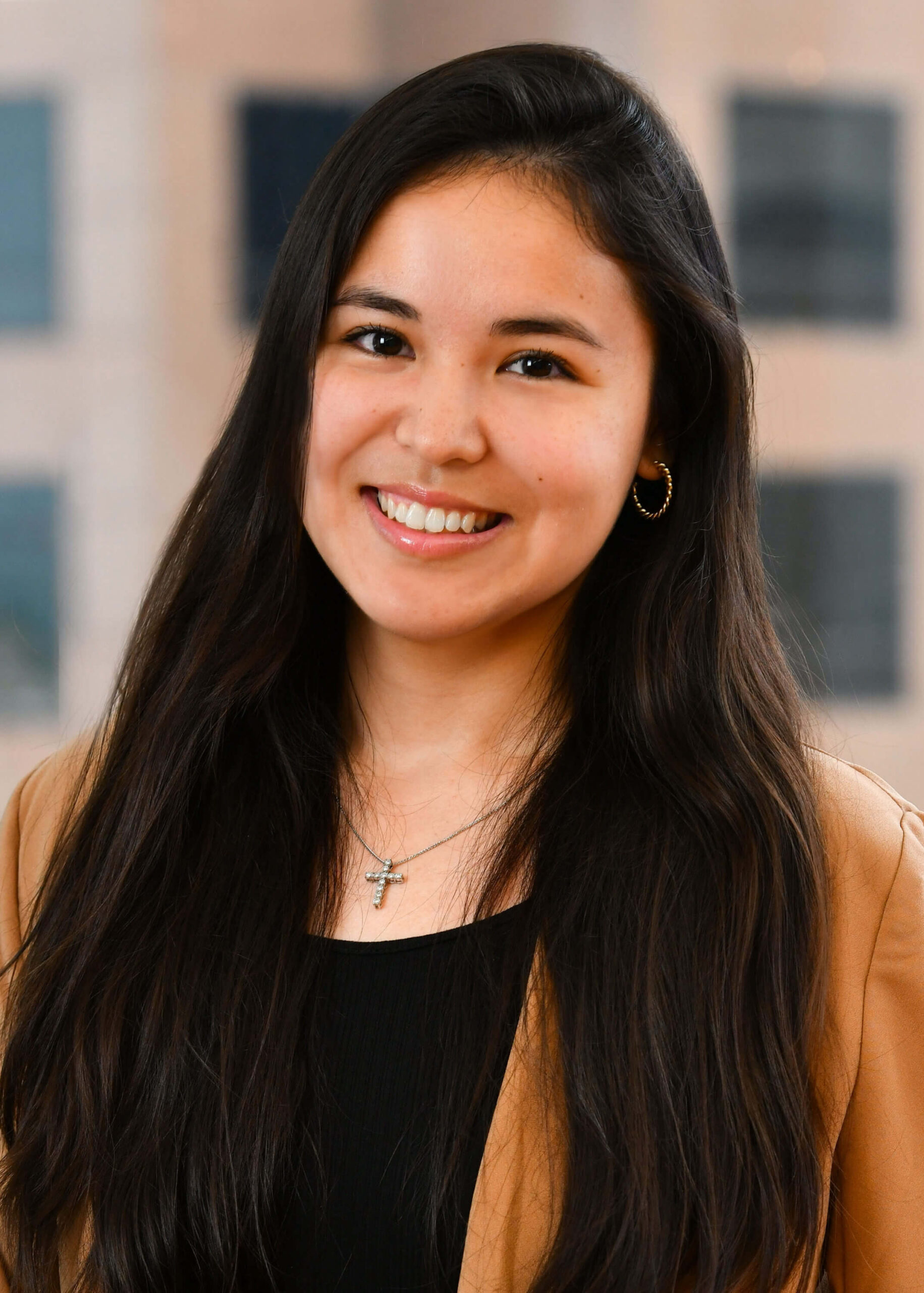 A woman with long dark hair smiles at the camera. She is wearing a black top, a tan blazer, gold hoop earrings, and a cross necklace. The blurred background shows tall windows and buildings.