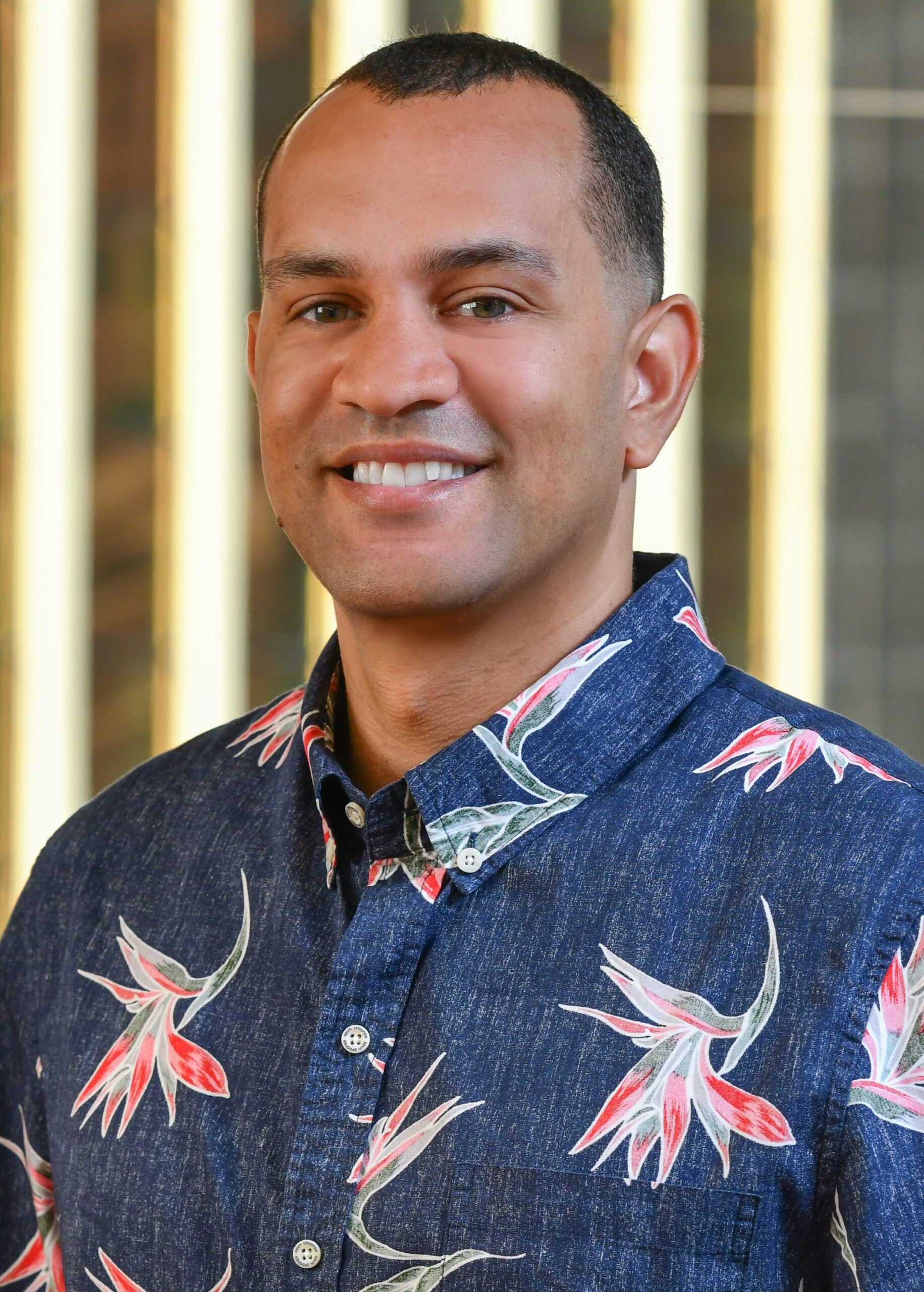A person smiles at the camera, wearing a blue button-up shirt with a colorful floral pattern. The background features vertical, blurred light stripes.